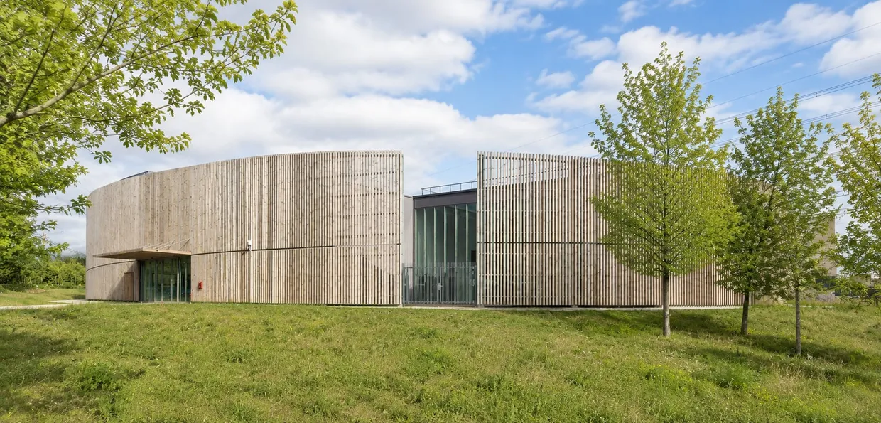 Le gymnase Cornuel vu du parc : façade courbe habillée de lattes de bois clair, entrée sous l'auvent, pelouse et jeunes arbres.
